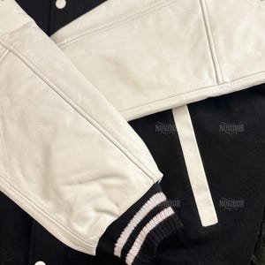 May include: Black and white varsity jacket with white leather sleeves. The jacket features a black body with white stripes and a ribbed knit cuff with white stripes. The jacket has white buttons.