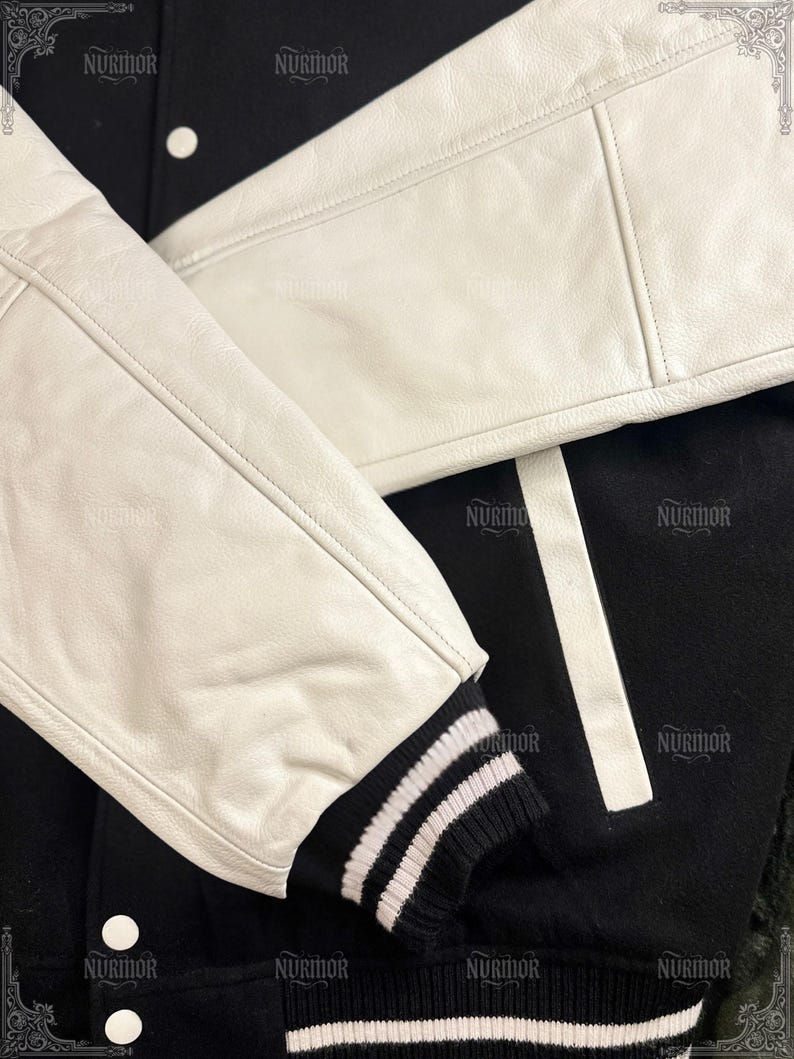 May include: Black and white varsity jacket with white leather sleeves. The jacket features a black body with white stripes and a ribbed knit cuff with white stripes. The jacket has white buttons.