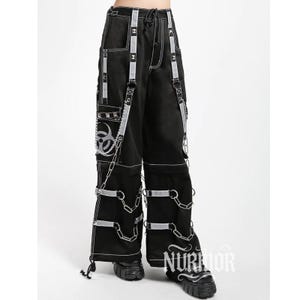 May include: Black wide-leg cargo pants featuring white contrast stitching, silver-toned metal chains, and reflective-style straps. The pants include a biohazard symbol graphic on the side pocket, multiple metal D-rings, and adjustable leg straps.