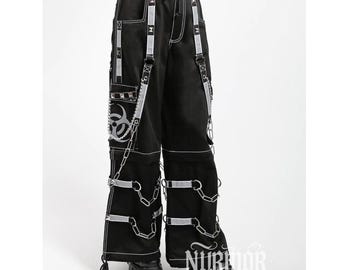 Könnte beinhalten: Schwarze Cargohose mit weitem Bein, weißen Kontrastnähten, silberfarbenen Metallketten und reflektierenden Riemen. Die Hose verfügt über ein Biohazard-Symbol auf der Seitentasche, mehrere D-Ringe aus Metall und verstellbare Beingurte.