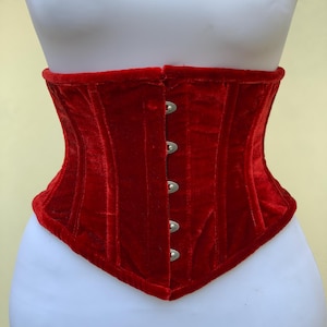 May include: A vibrant red velvet corset featuring a structured design with vertical boning channels and a front closure consisting of five silver-toned metal busk hooks. The garment has a curved hemline and a soft, plush texture throughout.