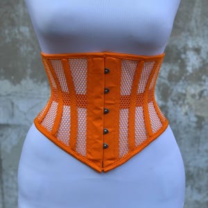 May include: An orange corset featuring vertical fabric panels alternating with sections of orange mesh. The garment has a front closure with silver-toned metal busk hooks and a pointed hemline, displayed on a white mannequin torso.