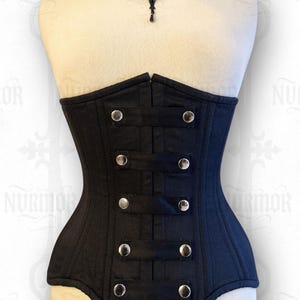 May include: A black corset with silver buttons and straps, shown on a mannequin. The corset has a structured design and a fitted shape. A black lace choker with a pendant is worn around the mannequin's neck. The corset is a piece of gothic fashion.