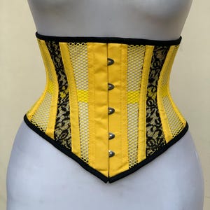 May include: A yellow and black corset featuring vertical panels of solid yellow fabric alternating with black lace and yellow mesh inserts. The garment has a front busk closure with five silver-toned metal clasps and black trim along the edges.