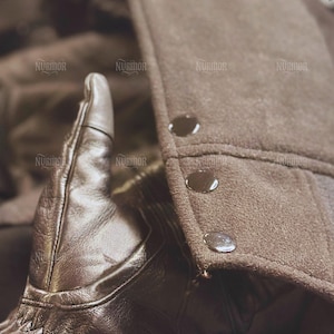 May include: Close-up of a brown leather glove with decorative stitching. The glove is next to a brown coat with three dark buttons. The coat has a textured, felt-like appearance.