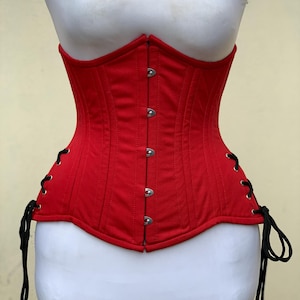 May include: A bright red corset featuring a front busk closure with five silver-toned metal clasps. The garment includes vertical panel stitching and black cord lacing on both sides, threaded through silver-toned metal grommets for a secure fit.