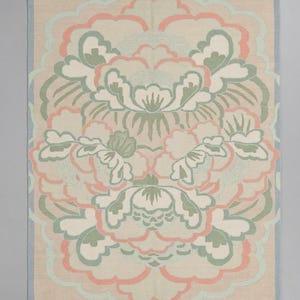 May include: A rectangular area rug featuring a symmetrical floral pattern in shades of sage green, soft peach, and cream on a beige background. The rug has a light blue border and short white fringe along the top and bottom edges of the textile.