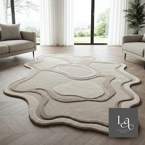 May include: An irregular, organic-shaped area rug in shades of beige and cream, featuring layered, wavy contours and a textured pile surface. A grey square logo in the bottom right corner displays the white text LA DESIGNER RUGS.