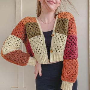 May include: A colorful, patchwork crochet cardigan in shades of orange, cream, brown, and burgundy. The open-front sweater features a V-neck, long sleeves, and wooden buttons. The crochet design has a geometric pattern.