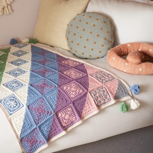 May include: A colorful, handmade crochet blanket with a geometric design. The blanket features squares in shades of green, blue, purple, and pink, with white accents. Tassels in coordinating colors adorn the corners. The blanket is draped over a couch.