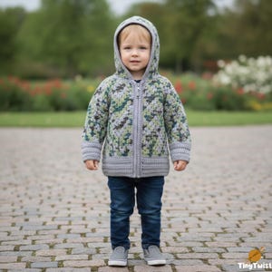 Könnte beinhalten: Ein Kind trägt eine handgefertigte Kapuzenjacke aus Häkelarbeit mit einem tarnähnlichen Muster in Grün-, Grau- und Brauntönen. Die Jacke hat einen durchgehenden Reißverschluss und wird mit einer blauen Jeans und grauen Turnschuhen kombiniert. Das Kind steht auf einem Ziegelboden.