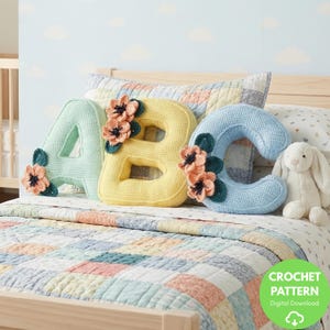 Op de afbeelding: Gehaakte letterkussens met de letters "ABC" in pastelkleuren, elk versierd met bloemaccenten. De "A" is mintgroen, de "B" is geel en de "C" is lichtblauw. Een wit pluchen konijn is ook zichtbaar. De afbeelding bevat een "CROCHET PATTERN" (haakpatroon) digitale download grafiek.