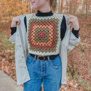 Pode incluir: Um colete de crochê feito à mão com um padrão de granny square. O colete apresenta um design quadrado com quadrados concêntricos em creme, verde-oliva, laranja e marrom. O colete é usado sobre uma gola alta preta e jeans azuis com um cinto preto.