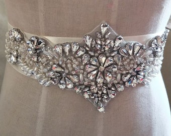 May include: A white satin bridal sash with a large, ornate crystal embellishment. The embellishment is made of clear rhinestones and pearls, arranged in a floral pattern.