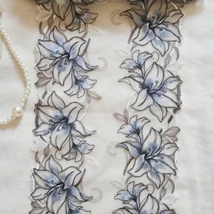May include: White lace trim with a floral design featuring blue and gray lilies. The trim is 24 inches wide.