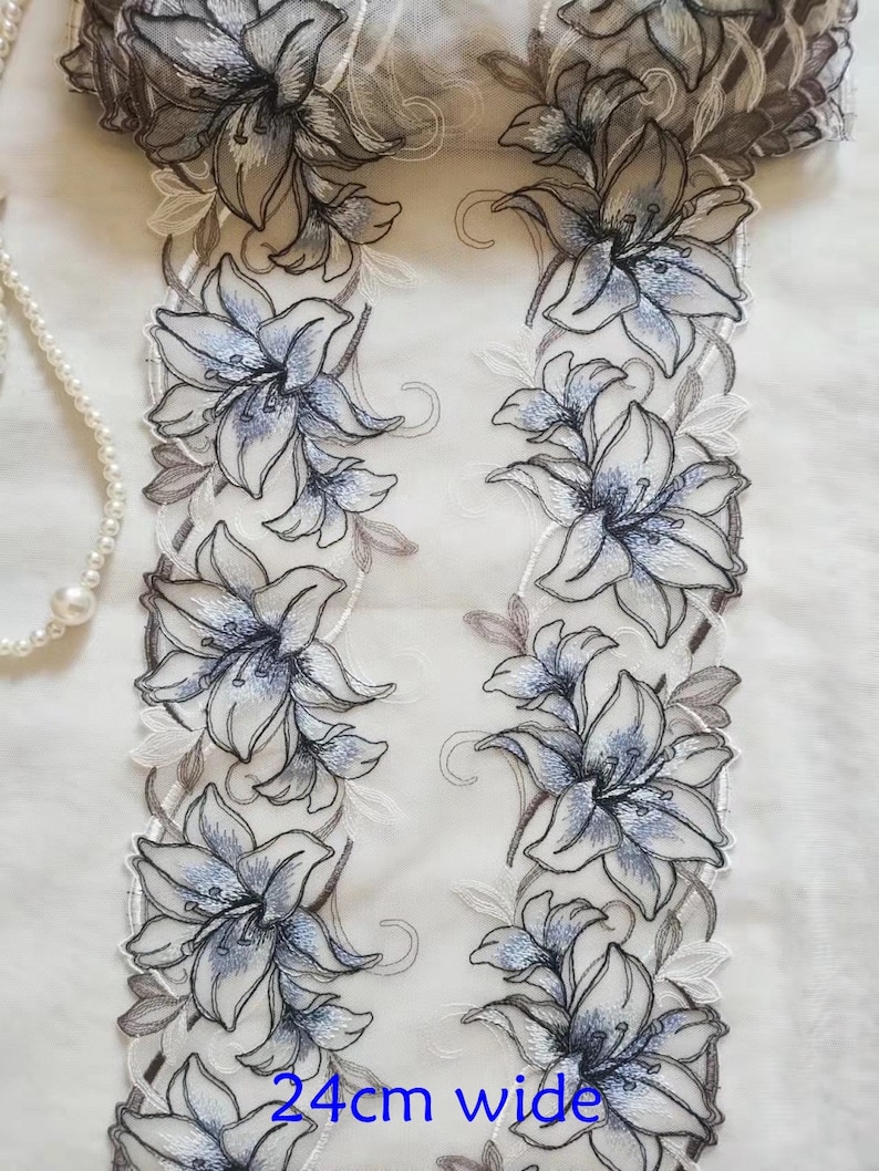 May include: White lace trim with a floral design featuring blue and gray lilies. The trim is 24 inches wide.