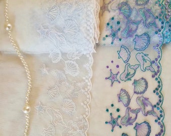 May include: Two white lace trims with a scalloped edge. The trim on the top has a design of seashells and mermaids. The trim on the bottom has a design of seashells, mermaids, and starfish in shades of purple, teal, and white.