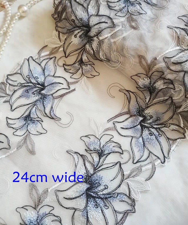 May include: A white sheer fabric with embroidered gray and blue lilies. The fabric is 24 inches wide.