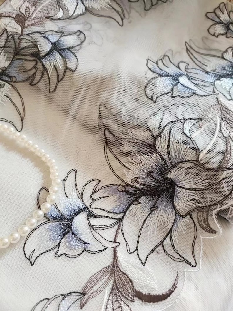 May include: White sheer fabric with embroidered floral designs in shades of gray and blue. The fabric has a delicate, airy feel and would be perfect for a variety of crafting projects.