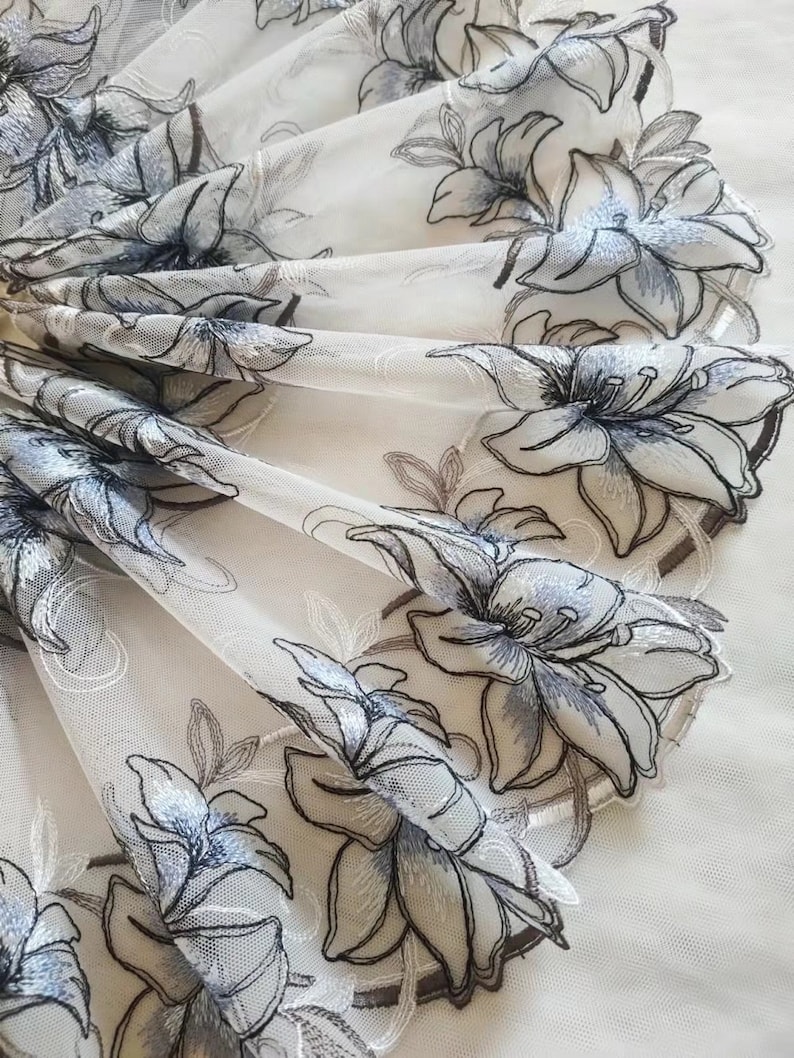 May include: White sheer fabric with a delicate floral embroidery design. The flowers are outlined in black and have a light blue center.