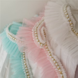 May include: Three layers of ruffled fabric trim with pearl beading. The top layer is white, the middle layer is pink, and the bottom layer is light blue.