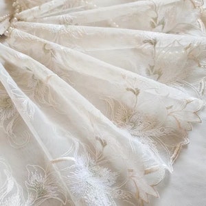 May include: White sheer fabric with delicate floral embroidery in shades of cream and green. The fabric has a scalloped edge.