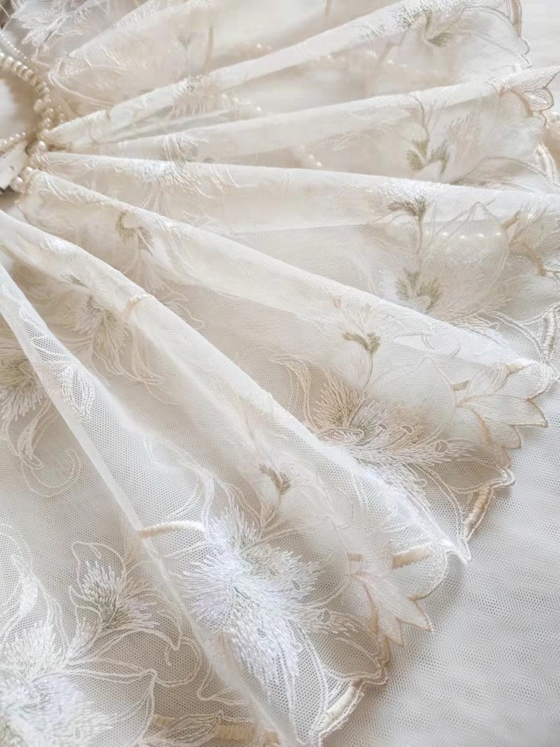 May include: White sheer fabric with delicate floral embroidery in shades of cream and green. The fabric has a scalloped edge.