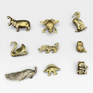 May include: A collection of nine small, gold-colored animal figurines. The assortment includes a hippo, frog, swan, dragon, owl, peacock, stegosaurus, and a bear holding leaves. Each figurine is detailed and has a metallic finish.