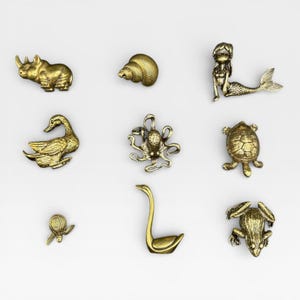 May include: A collection of nine small, gold-colored animal figurines. The figurines include a rhinoceros, seashell, mermaid, duck, octopus, turtle, snail, swan, and frog. Each figurine is highly detailed and has a metallic finish.