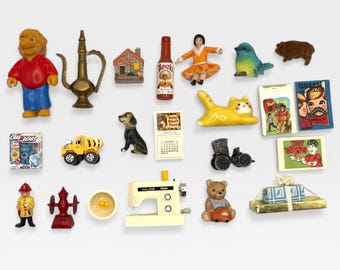 May include: A collection of miniature figurines and objects, including a Care Bears box, a yellow truck, a black dog, a calendar, a yellow cat, a sewing machine, a teddy bear, and small books. The items are arranged on a light background.