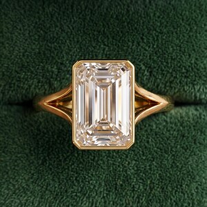 May include: A gold ring featuring a large, rectangular emerald-cut clear gemstone set in a bezel setting. The band has a split shank design that tapers towards the centre stone. The ring is displayed inside a dark green velvet-lined wooden box.