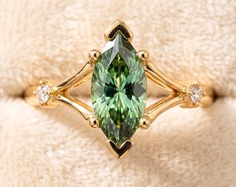 May include: A gold ring with a marquise-cut green gemstone as the centerpiece. The ring features two small, round, clear gemstones on either side of the main stone. The ring is set in a gold band with a delicate, openwork design. The ring is displayed in a beige ring box.