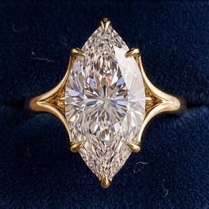 Op de afbeelding: Een ring met een heldere edelsteen in marquise-slijpvorm, gezet in een band van gepolijst geelgoud. De steen heeft een gefacetteerd oppervlak in een puntige ovale vorm, vastgezet door zes klauwen die uit de gespleten metalen band komen.