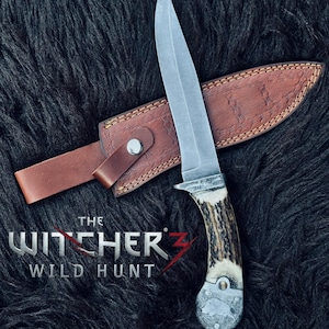 May include: A hunting knife with a silver blade and a handle made of antler material. The knife is in a brown leather sheath. The text "THE WITCHER 3 WILD HUNT" is visible.