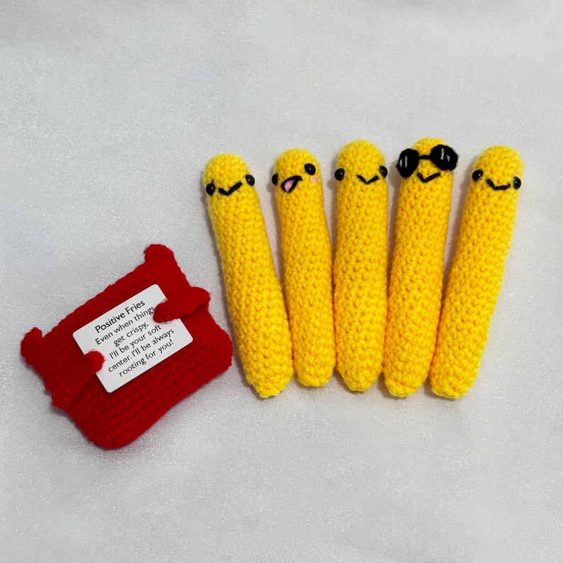 May include: A collection of five yellow crochet french fries with smiling faces, one wearing sunglasses, next to a red crochet container. The container has a tag that reads "Positive Fries".