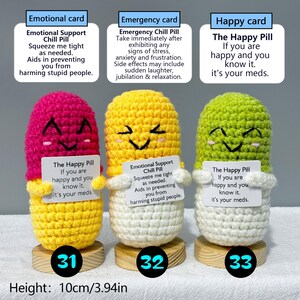 May include: Three handmade crochet pill-shaped toys in pink, yellow, and green, each with a smiling face and a small card. The cards have different messages. The height of each toy is 3.94 inches. Each toy is on a small wooden base.