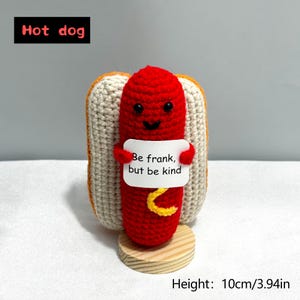 May include: A handmade, crocheted hot dog with a smiling red sausage, held in a beige bun. The hot dog holds a sign that reads "Be frank, but be kind." The height is 3.94 inches.