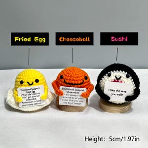 May include: Three crocheted food-themed figurines: a yellow fried egg, an orange cheeseball, and a black and white sushi roll. Each has a smiling face and holds a sign with text. The height is 1.97 inches.