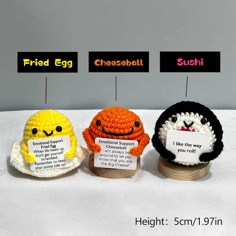 May include: Three crocheted food-themed figurines: a yellow fried egg, an orange cheeseball, and a black and white sushi roll. Each has a smiling face and holds a sign with text. The height is 1.97 inches.