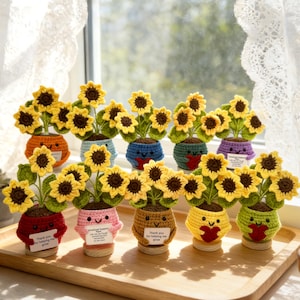 May include: A display of crocheted sunflower plants in different coloured pots, each with a smiling face. The sunflowers have yellow petals and brown centres. Some pots have small signs with text. The plants are on a wooden tray.