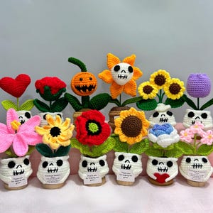 May include: A collection of crocheted flowers and plants, each potted in a white skull-shaped base with black facial features. The plants include a red heart, a red rose, a jack-o'-lantern, a skull flower, sunflowers, a tulip, and pink lilies.