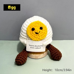 May include: A crocheted egg-shaped plush toy with a smiling yellow yolk and white egg white. The toy has brown legs and a small sign that reads "You're absolutely EGG-cellent!" The height is 3.94 inches.