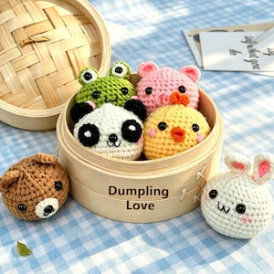 May include: A bamboo steamer basket labelled Dumpling Love containing four crocheted animal plushies: a green frog, a pink pig, a white panda, and a yellow chick. Two additional crocheted animal plushies, a brown bear and a white rabbit, sit outside.