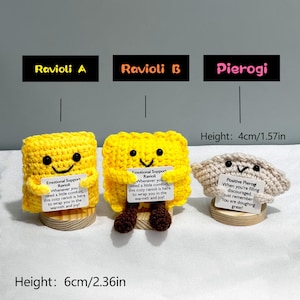 May include: Three handmade crochet food items: two yellow ravioli and one beige pierogi. The ravioli have smiling faces and the text "Emotional Support Ravioli." The pierogi has a smiling face and the text "Positive Pierogi." The ravioli are 6cm/2.36in tall, and the pierogi is 4cm/1.57in tall.