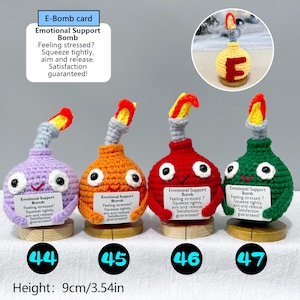 May include: Four handmade crochet "Emotional Support Bombs" in purple, orange, red, and green, each with a smiling face and a flame-like top. Each bomb is approximately 3.54 inches tall and includes a small wooden base.