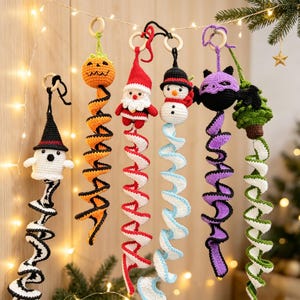 May include: Six crocheted hanging ornaments featuring a ghost, pumpkin, Santa Claus, snowman, bat, and Christmas tree. Each figure is attached to a colorful spiral tail and a wooden ring. The ornaments are crafted with yarn in various colors.