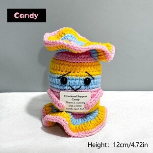May include: A handmade crochet candy figure in yellow, pink, and blue. The figure has a smiling face with black eyes and a small sign that reads "Emotional Support Candy." The height is 4.72 inches.