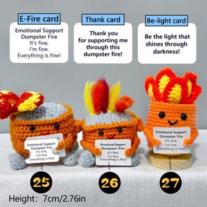 May include: Three crocheted figures resembling flames, each with a unique design and expression. They hold cards with humorous phrases like "Emotional Support Dumpster Fire." The height is 2.76 inches.