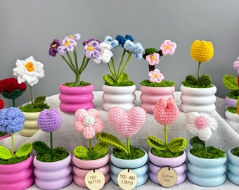May include: A collection of colorful crochet flowers in small, decorative pots. The flowers include a variety of styles, such as tulips, daisies, and heart-shaped designs. Some pots have wooden tags with phrases like "You Matter" and "You Are Loved".