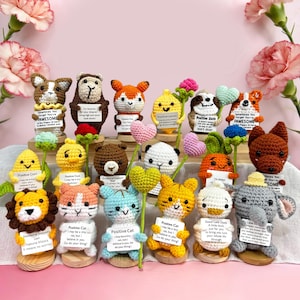 Puede incluir: Una colección de coloridas figuras de animales de ganchillo hechas a mano, cada una con un pequeño letrero con afirmaciones positivas. El surtido incluye un perro, un oso, un zorro, un limón, un perezoso y más, todo sobre un fondo floral rosa.